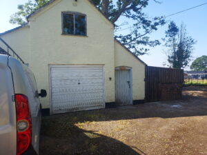 Exterior of the Mister Micawbers Cottage Doors workshop at Grindley Cottage, Crewe, a cream painted brick building with a white garage door surrounded by trees